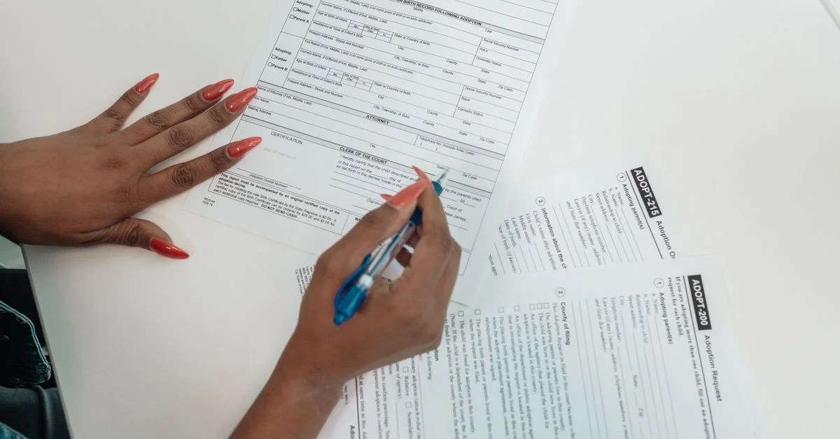 Une femme aux ongles rouges remplit des formulaires de demande d'adoption sur un bureau.