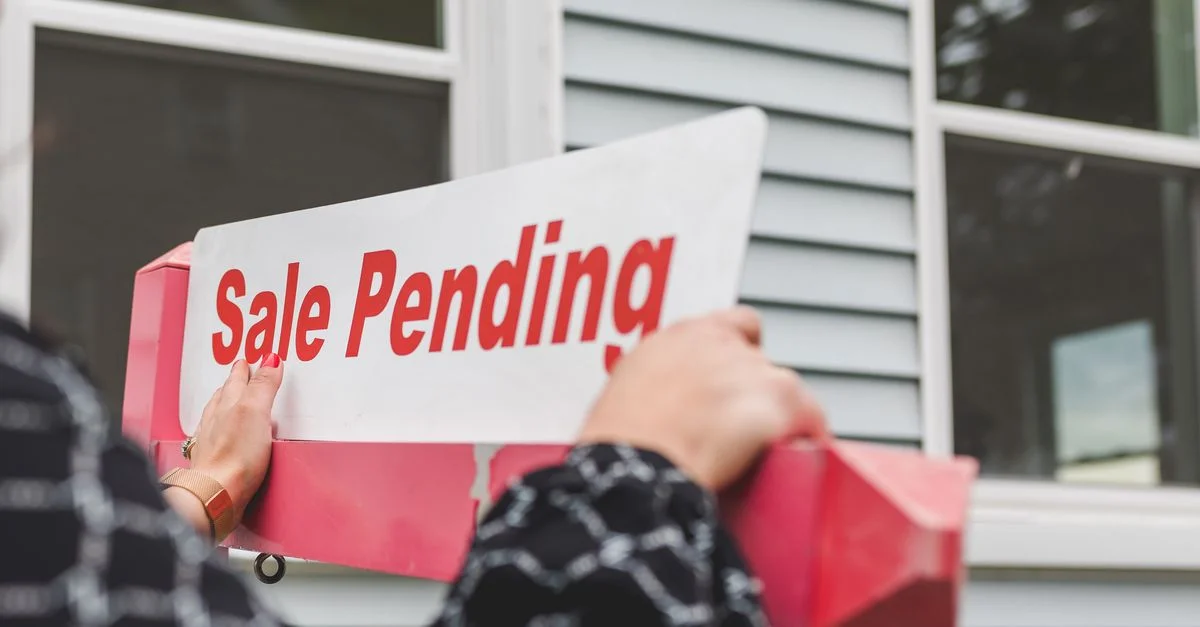 Gros plan de mains tenant un panneau 'Vente en cours' devant une maison, avec des thèmes immobiliers.